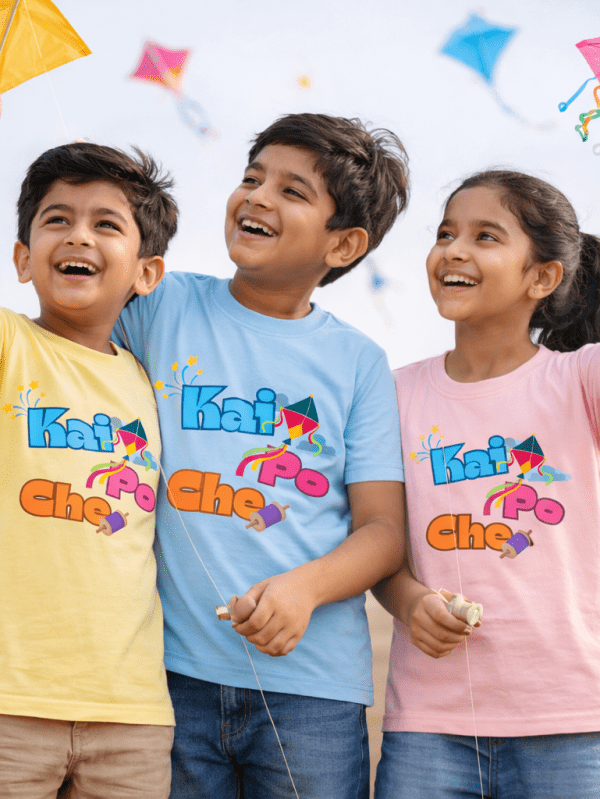Three Indian kids wearing Kai Po Che kite flying themed T-shirts, smiling and flying colorful kites outdoors